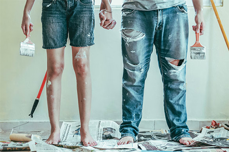 Couple wearing denim standing next to each other and holding paint brushes while doing DIY home improvement projects
